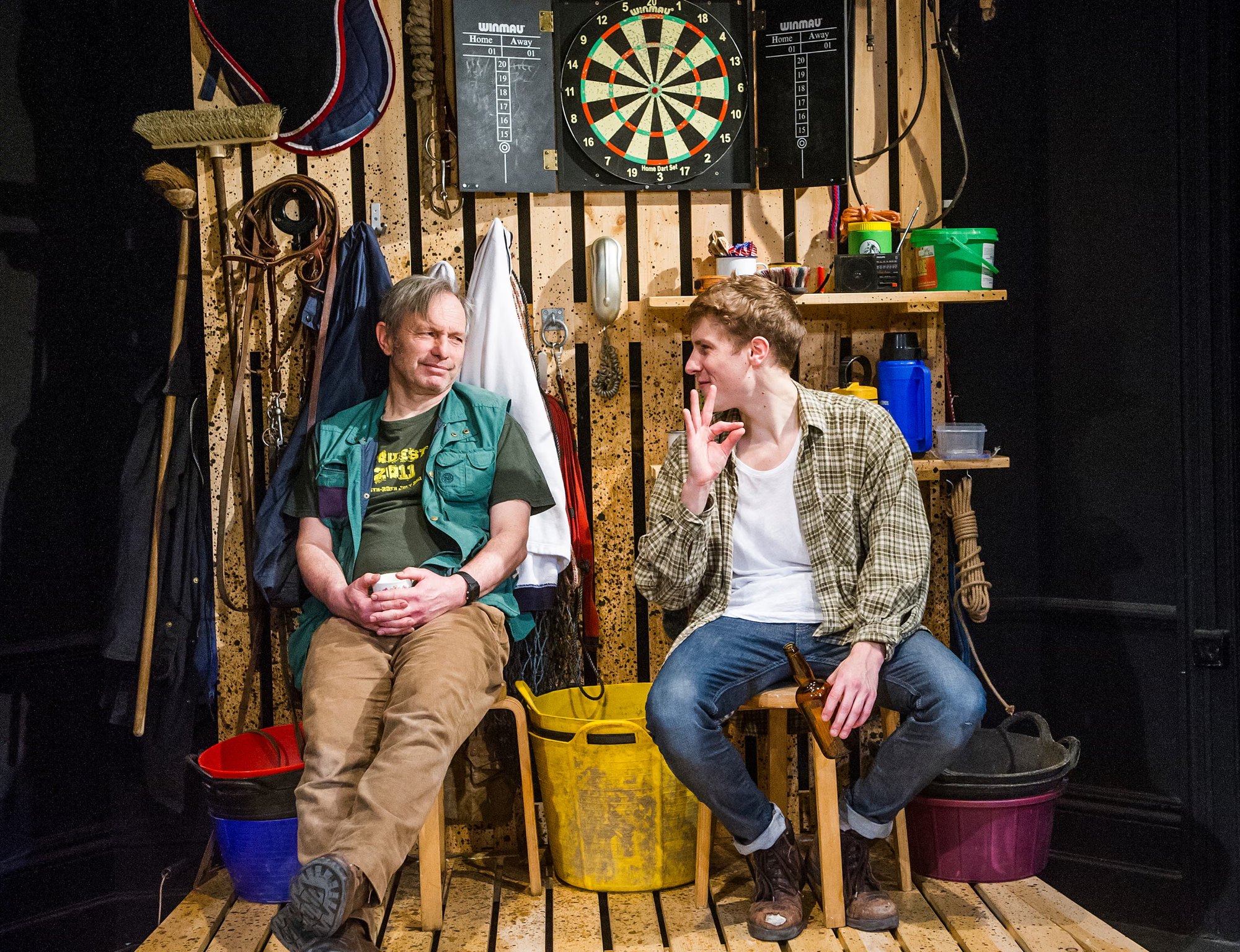Two men sit talking on stools in a shed-like set with a dartboard, tools and buckets; the younger one makes an okay sign with his hand