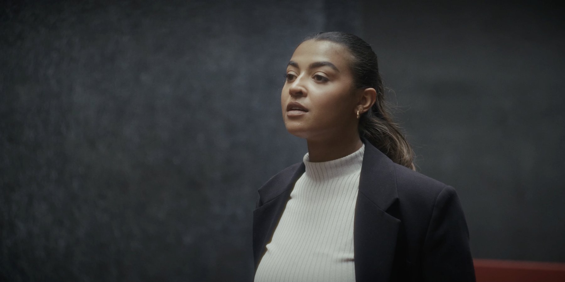Close-up of a woman in a dark blazer looking off-frame with parted lips, against a dark grey wall