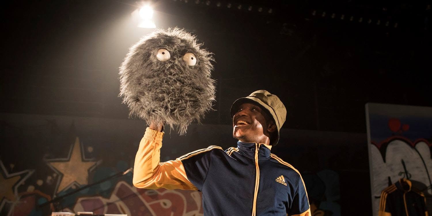 A performer in a bucket hat and tracksuit smiles up at a shaggy grey puppet held in the air, with boomboxes and graffiti behind