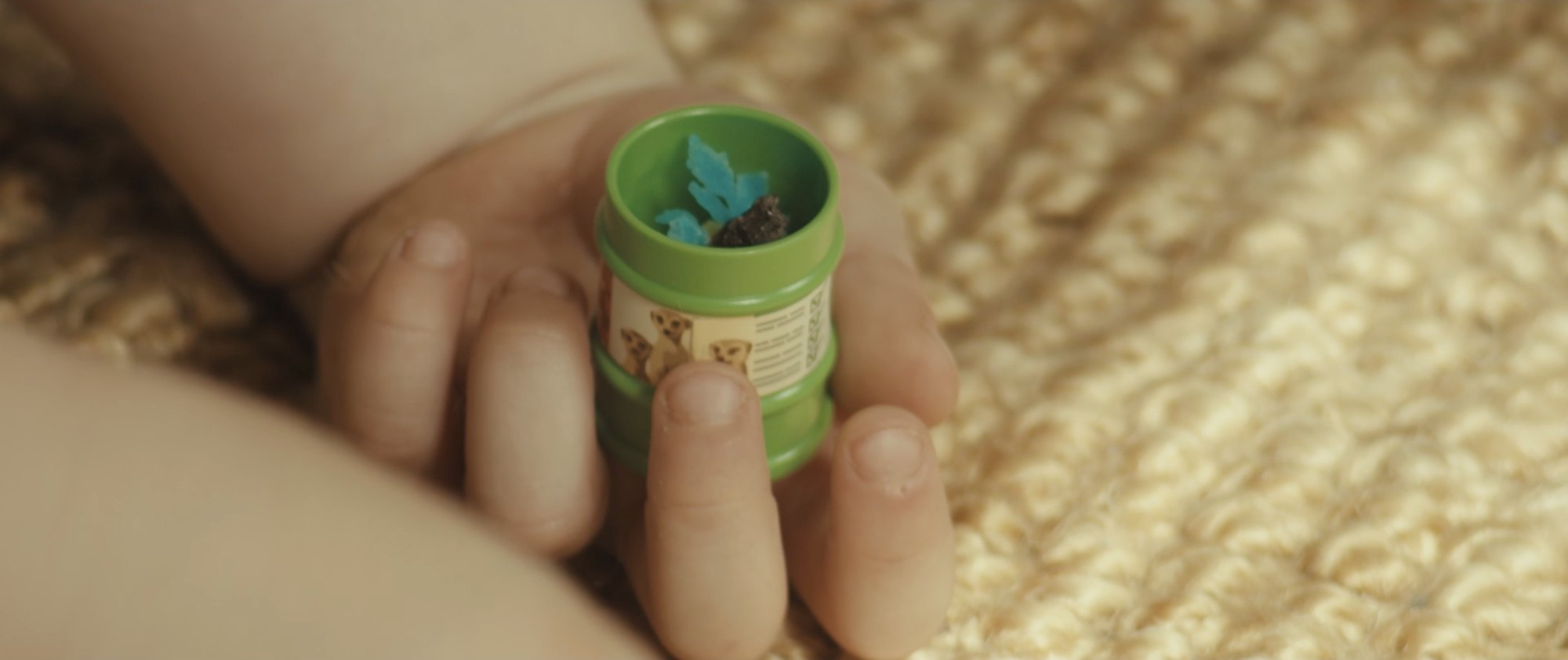 Film still from They’re Taking The Tiles Off The Building: a child’s hand holding a small green tub with a blue leaf inside