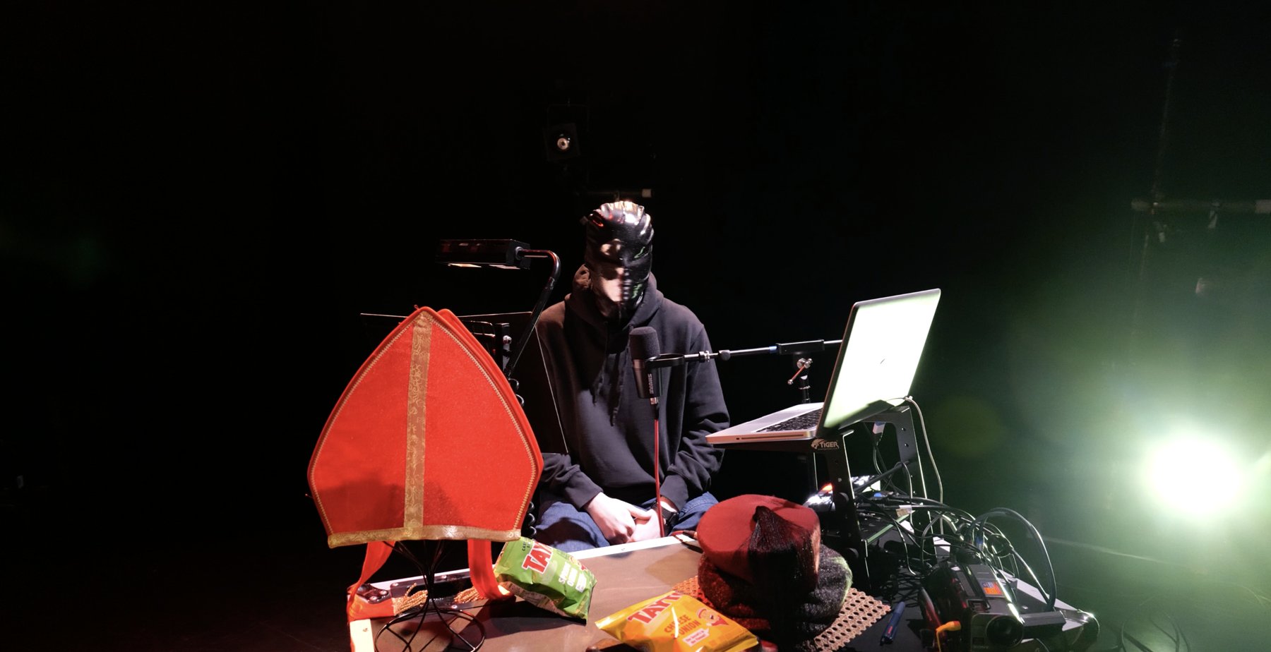 Performer wearing a black inflatable mask, seated at a table of props including a red mitre, crisps, a laptop and a camcorder