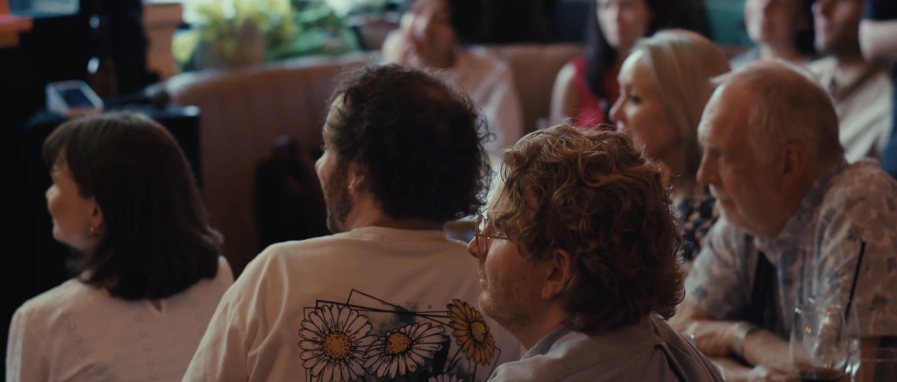 Film still: audience members seen from behind, listening at a reading
