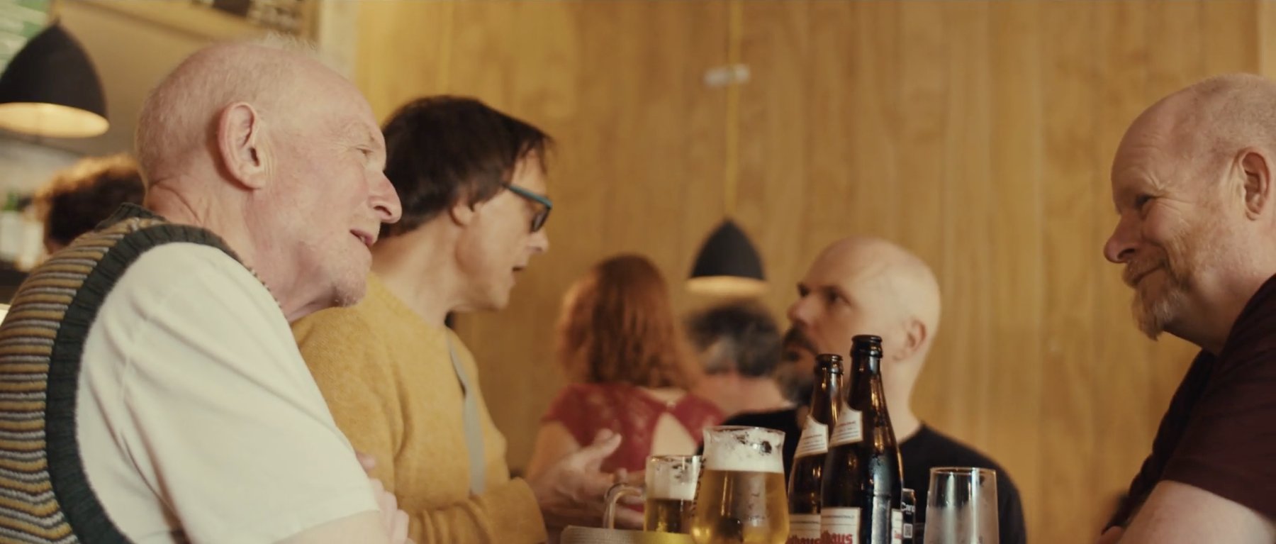 Film still: friends talking over drinks in a warm, wood-panelled bar