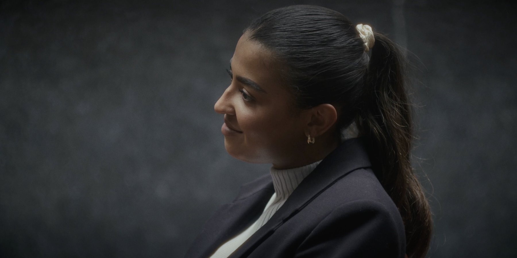 Profile of a woman with her hair in a ponytail and a cream scrunchie, softly lit against a dark grey backdrop