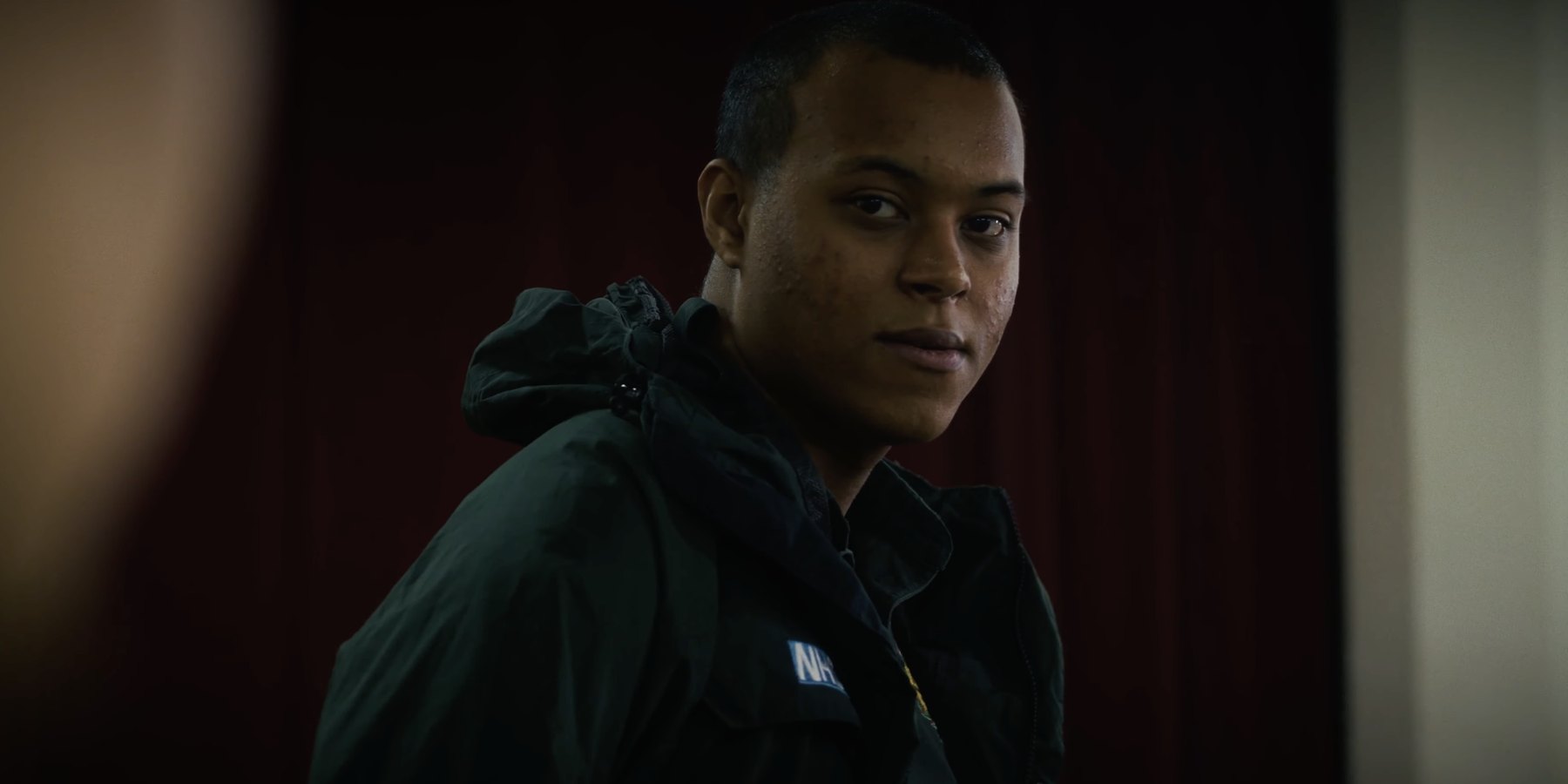 Film still from HORIZON: a young man in a dark ambulance jacket glances towards the camera in front of a red curtain