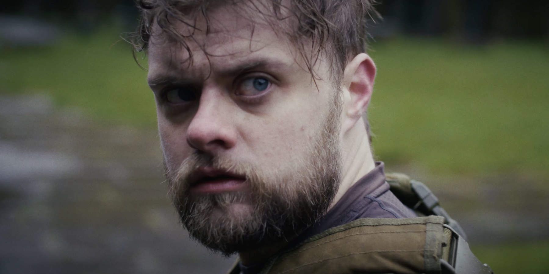 Film still from HORIZON: close-up of a bearded man with wet hair glancing back over his shoulder, in a tactical vest on a grassy field