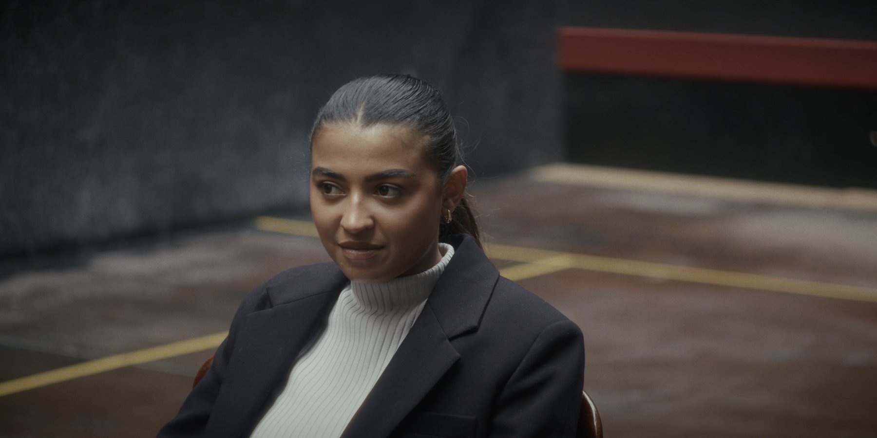 A woman seated in a blazer glances to one side with a faint smile, in a dim hall with yellow floor markings