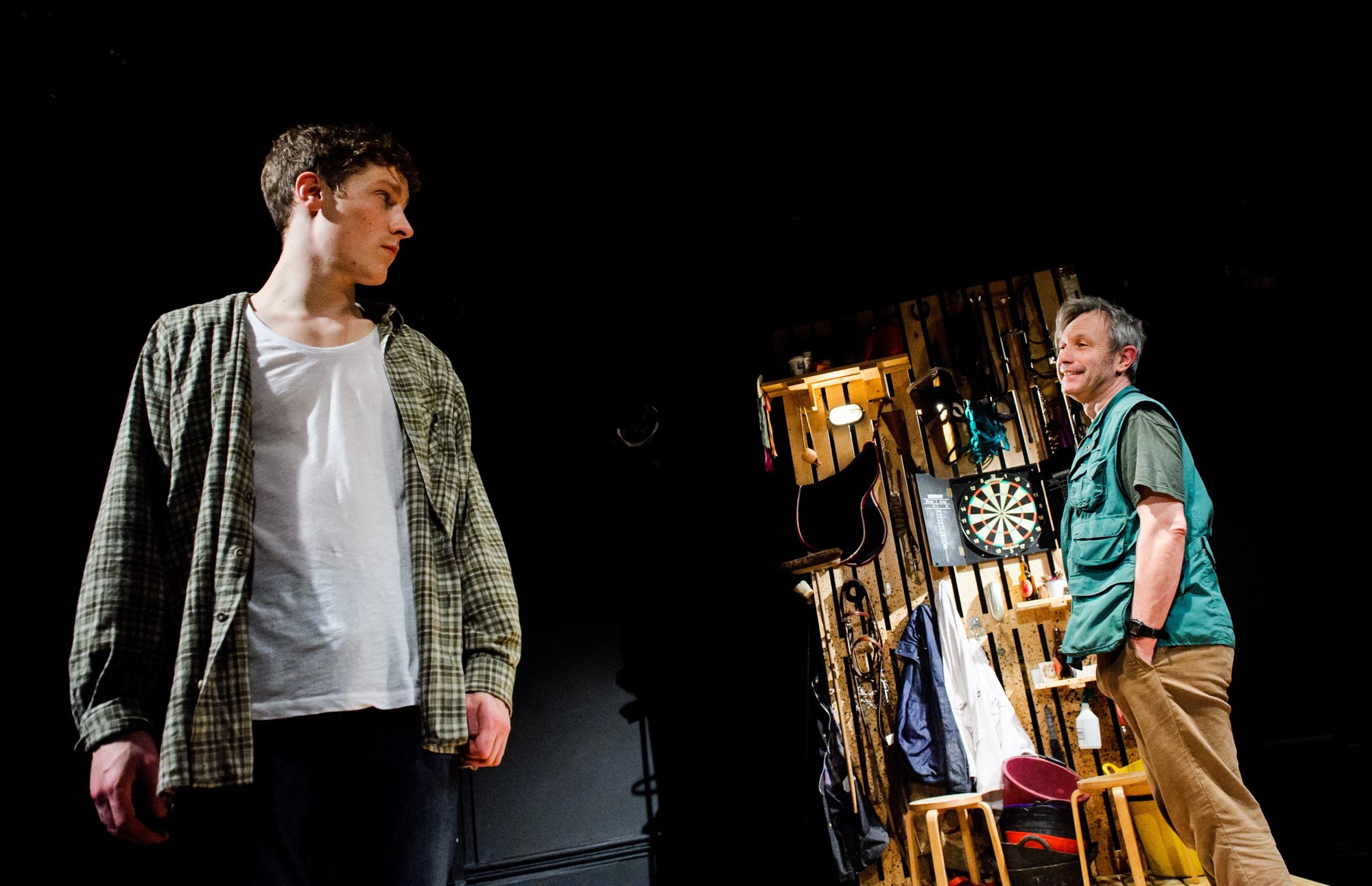 Two actors on a dark stage: a young man in a checked shirt looks across at an older man in a green vest beside a wall of tools and a dartboard