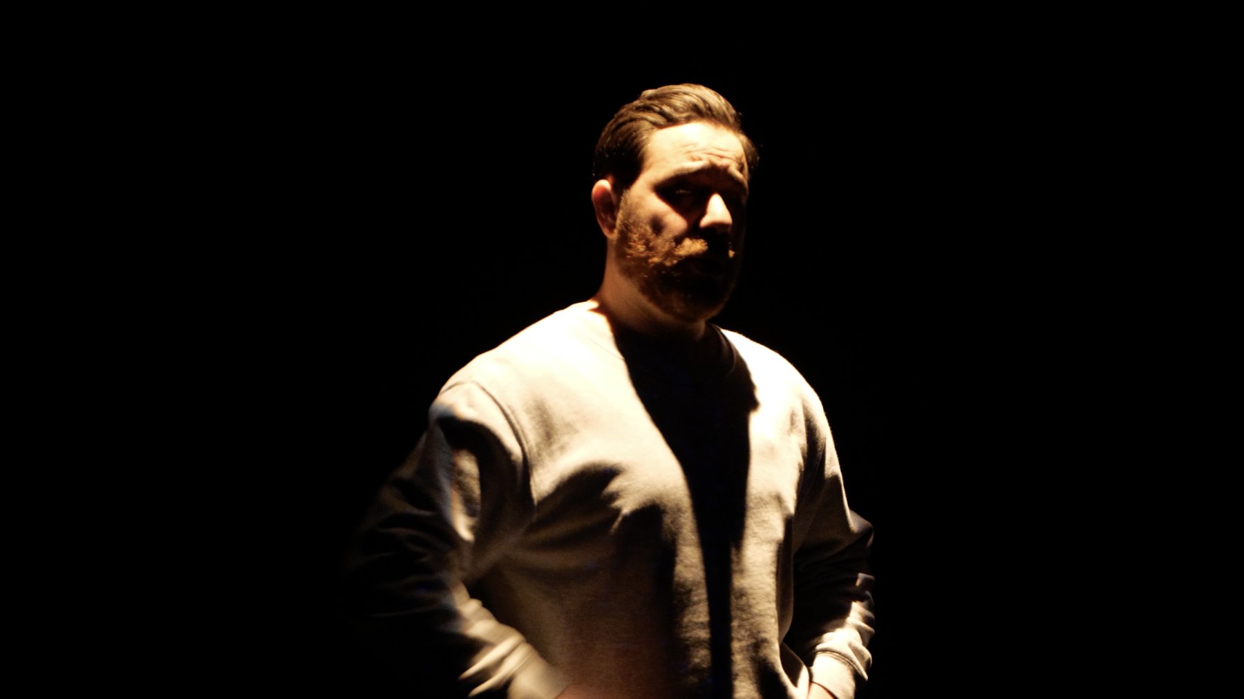 Close portrait of a bearded performer in a grey sweatshirt, lit from the side against black