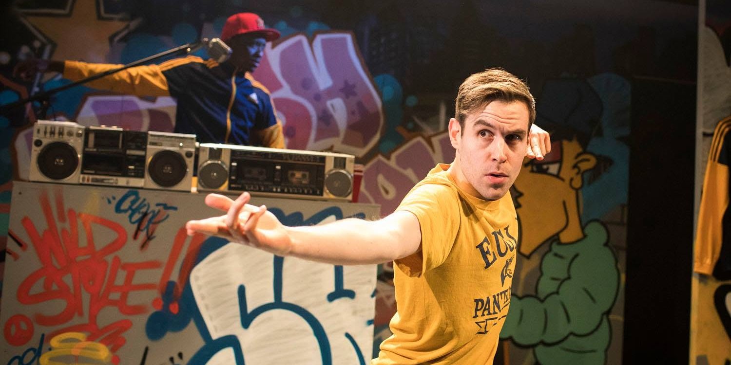 A performer in a yellow T-shirt lunges with an outstretched arm in front of a graffiti wall and boomboxes, with another performer in a red cap behind
