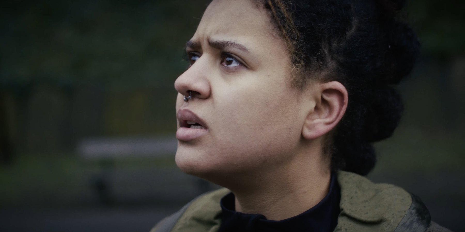 Film still from HORIZON: close-up of a woman with a septum ring looking up and off-frame, outdoors