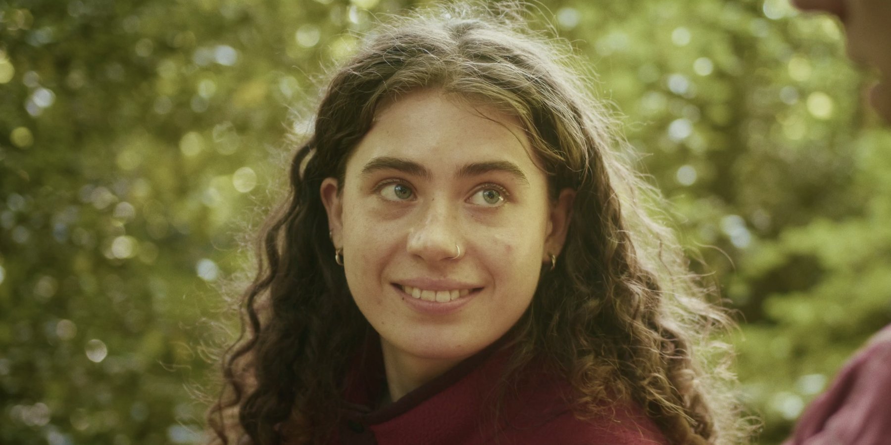 A young woman with long curly hair smiles and glances upward, with green foliage blurred behind her