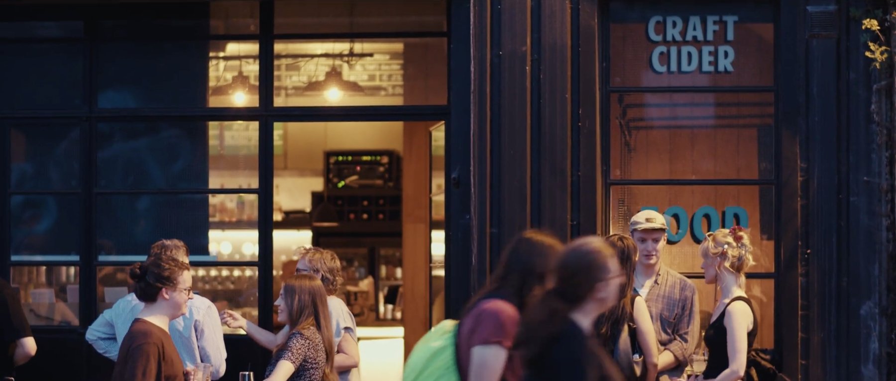 Film still: people chatting outside a bar at dusk