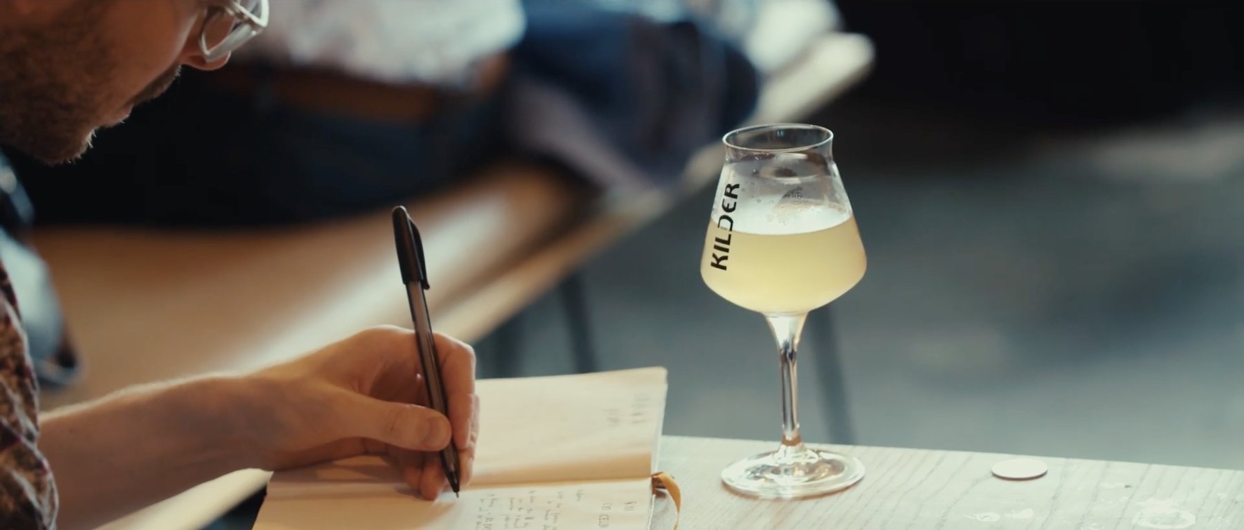Film still: a person writing in a notebook beside a glass of beer