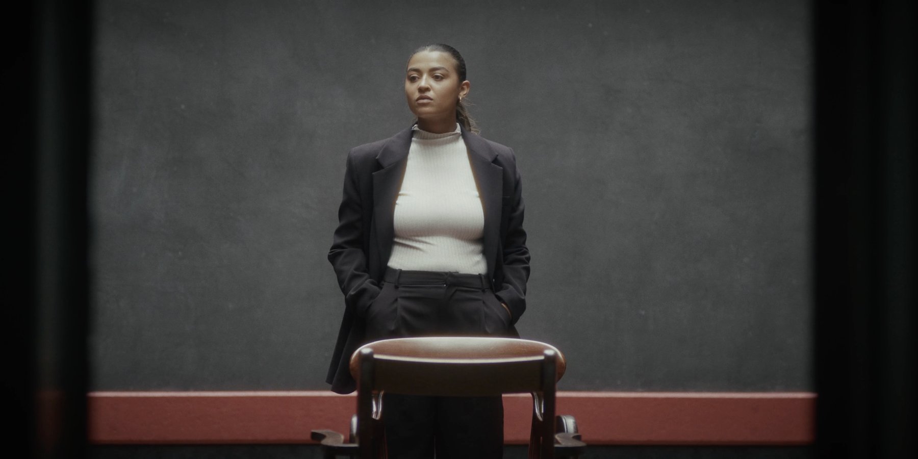 A woman in a dark blazer and white top stands with her hands in her pockets behind an empty wooden chair, against a grey wall