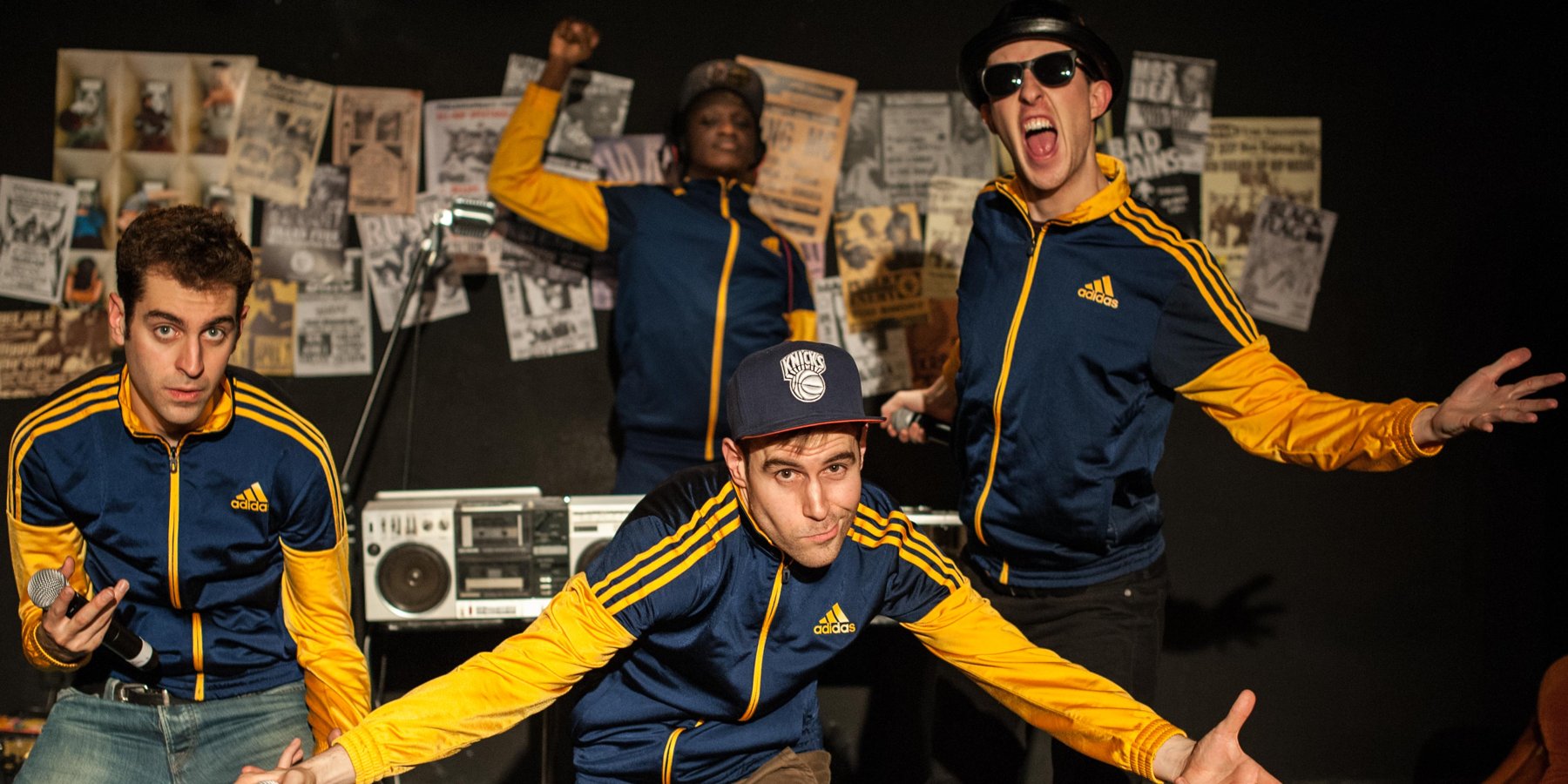 Four performers in matching navy and yellow tracksuits strike poses in front of a wall of old gig flyers and a boombox