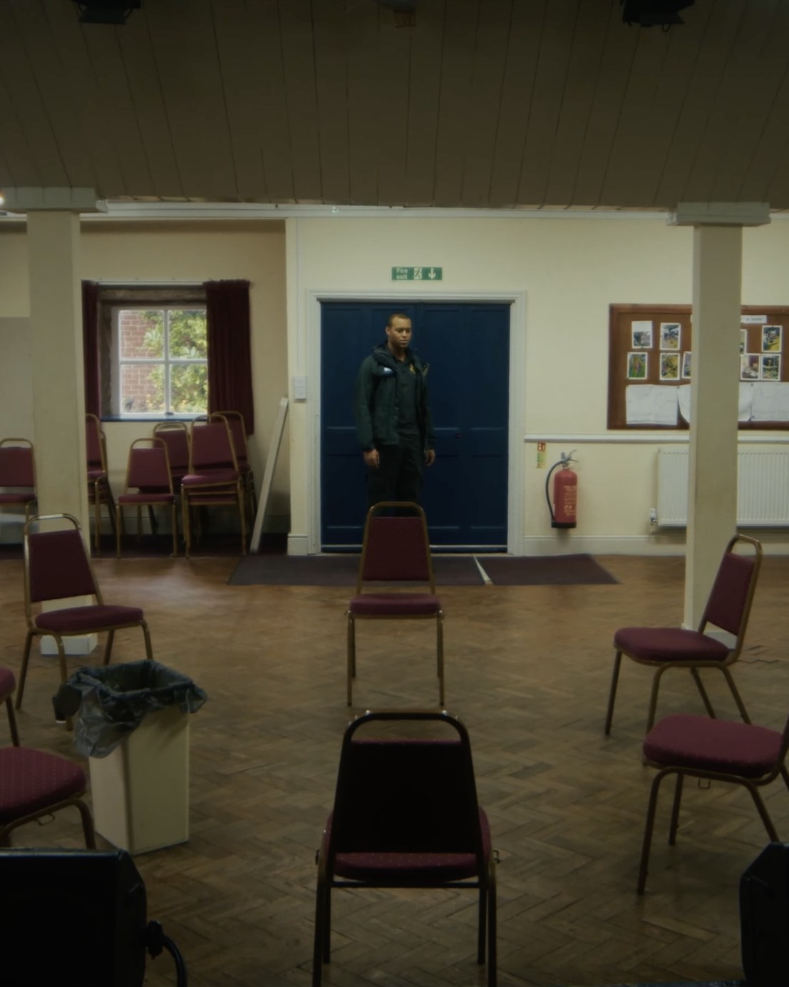 Film still from HORIZON: an empty community hall with red chairs set out in a circle on a parquet floor, a lone figure standing at the far double doors