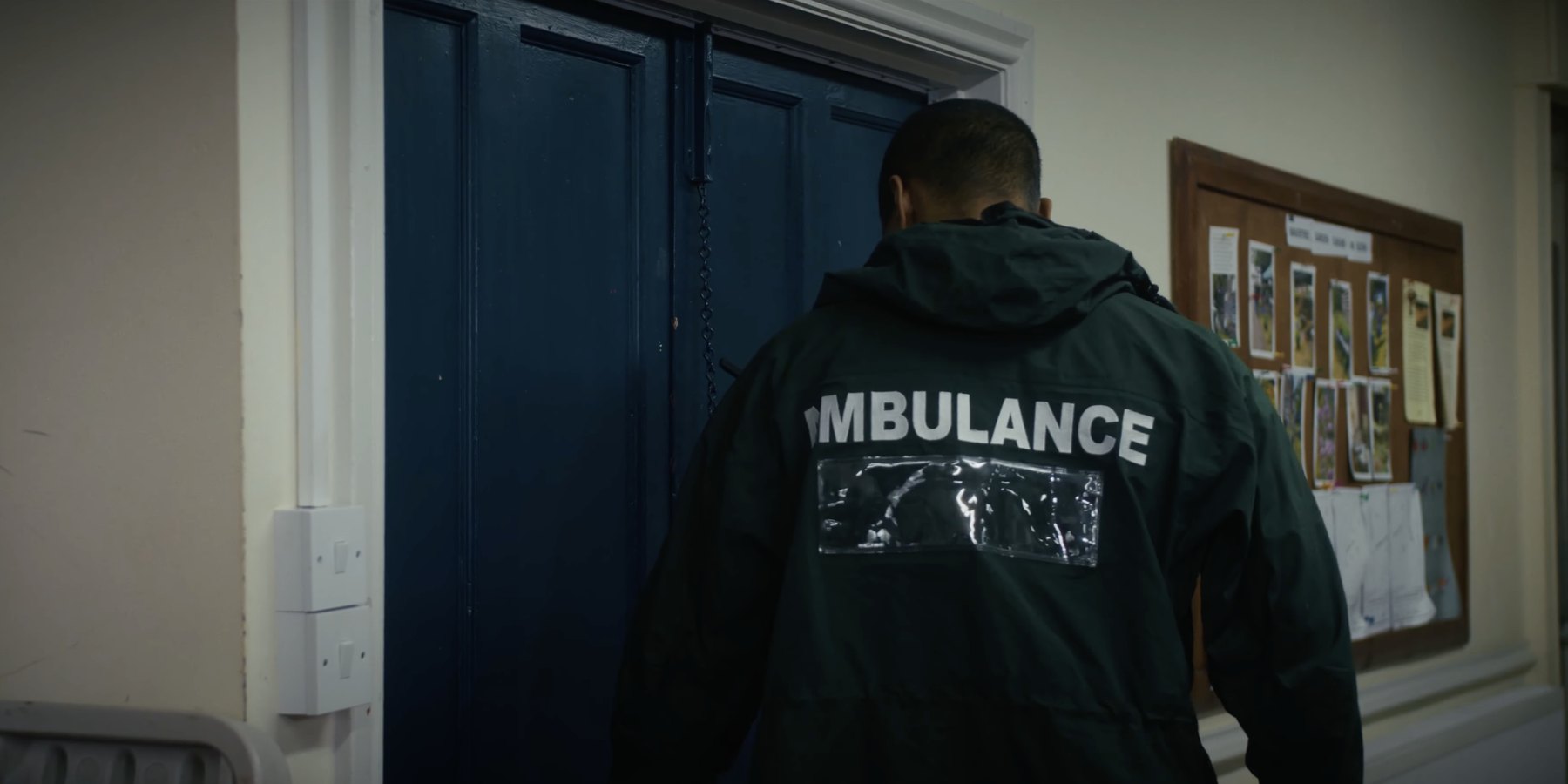 Film still from HORIZON: a man in a dark green ambulance jacket seen from behind, standing at a pair of blue double doors beside a noticeboard