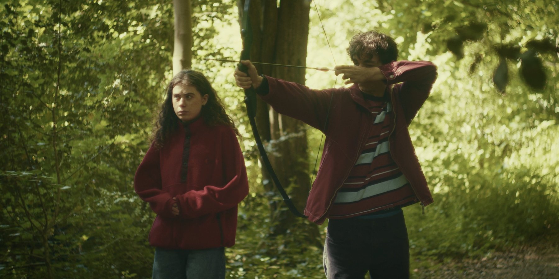 A young man draws a bow beside a young woman who watches something off-screen, in dappled green woodland