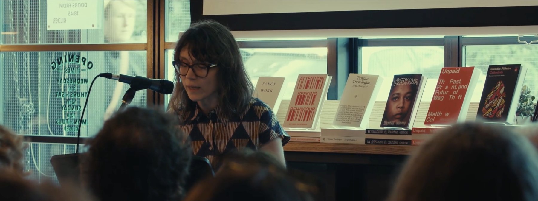 Film still: a woman in glasses reading at a microphone, with a shelf of books behind her