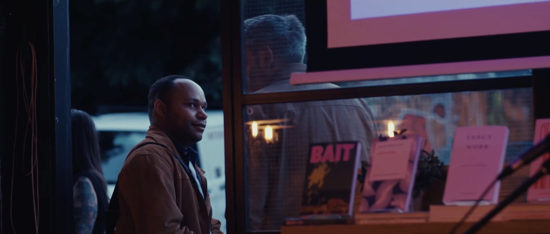 Film still: a man in a brown jacket glancing aside beside a window onto a book display