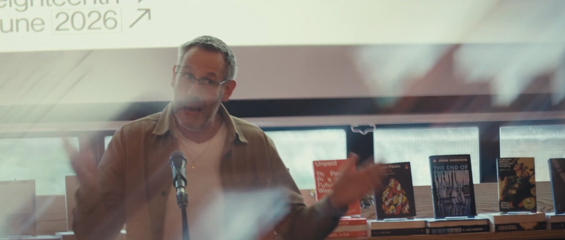 Film still: a man speaking at a microphone and gesturing, books on a shelf behind him
