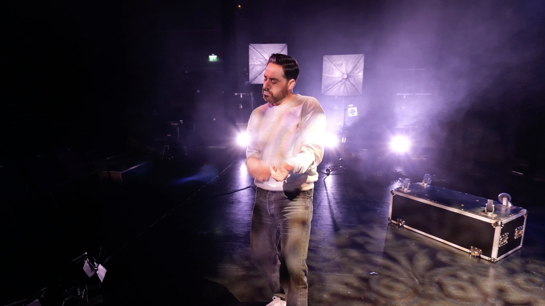 Performer in a grey sweatshirt on a hazy stage, backlit in purple, with a flight case and softboxes behind him
