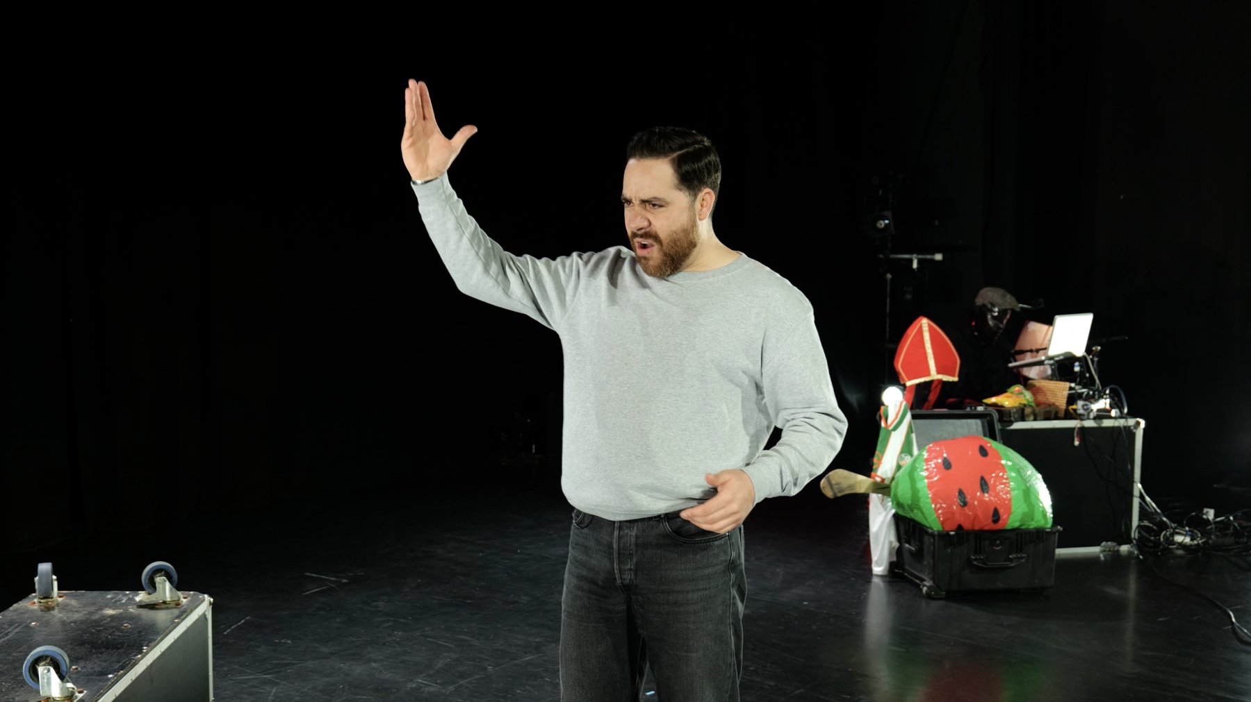 Performer mid-gesture with one arm raised, beside a flight case and an inflatable watermelon